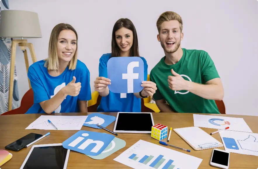 Three young professionals holding social media icons for Facebook, Twitter, and LinkedIn, discussing social media marketing strategies at a table with charts and devices, representing why social media marketing is essential for businesses in Kerala and Kochi.